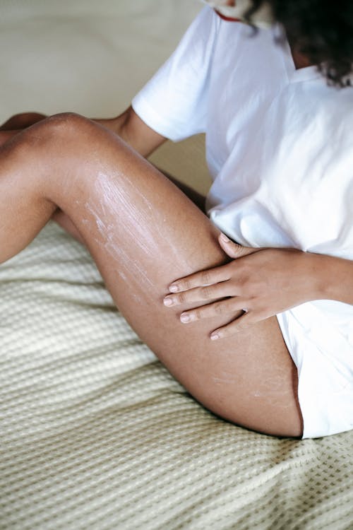 High angle  of unrecognizable female in white t shirt and with bared legs sitting on bed and applying cosmetic cream on leg