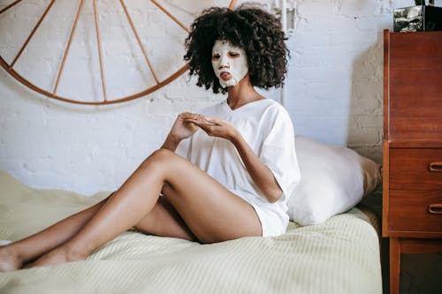 Young ethnic female with curly hair and with cosmetic mask on face sitting on bed and doing skincare routine in bedroom