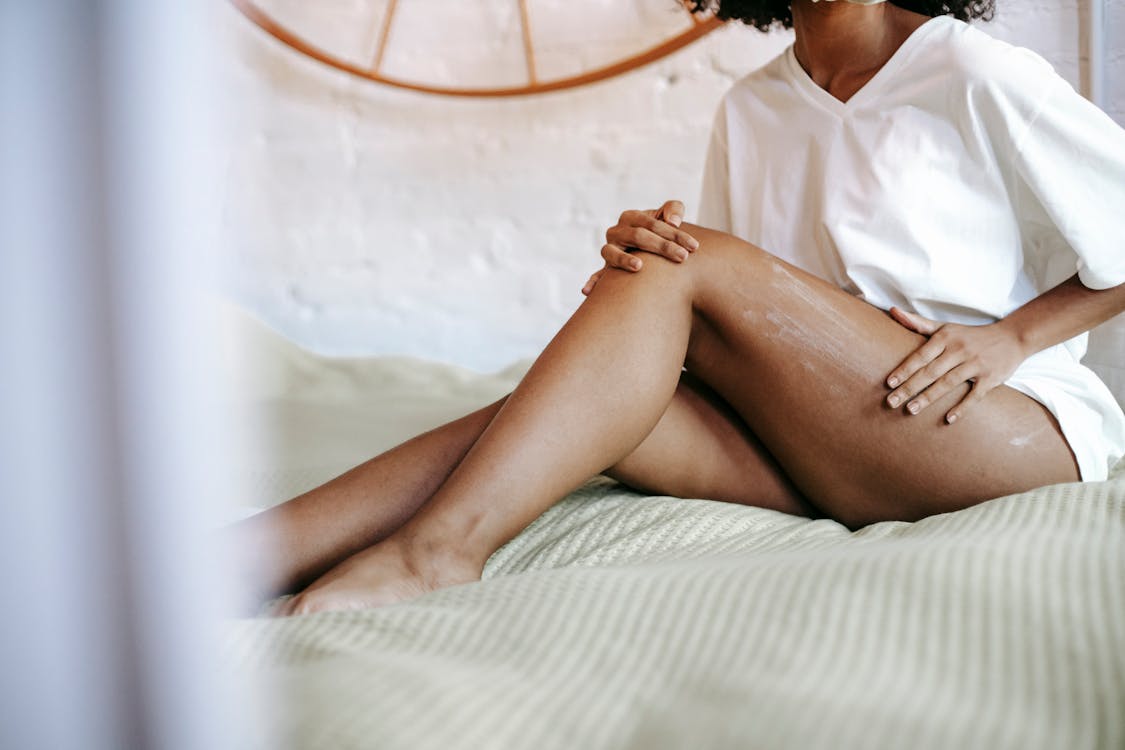 Free Side view of unrecognizable female in white t shirt sitting on bed and applying lotion on leg while resting in weekend Stock Photo