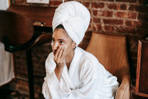 Free Young female in white bathrobe and towel turban applying eye patches on face while sitting at home Stock Photo