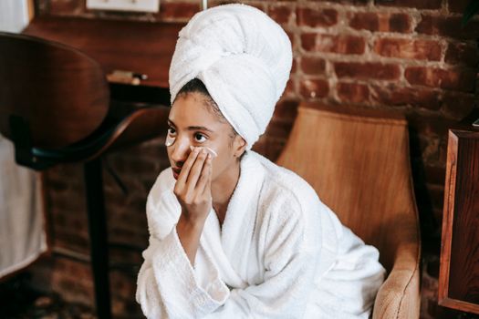 Young female in white bathrobe and towel turban applying eye patches on face while sitting at home