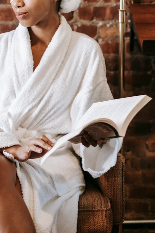 Free Young black  female in bathrobe and towel sitting on chair with book and enjoying daily routine while relaxing in cozy room Stock Photo