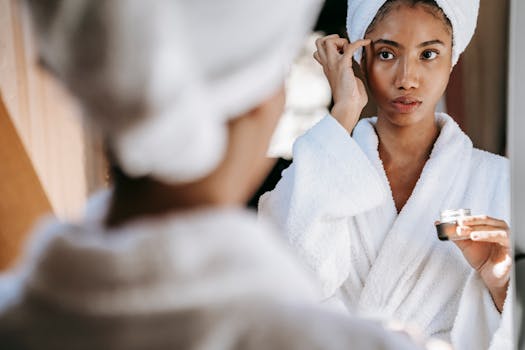 Young woman in bathrobe applying skincare cream while looking in the mirror for morning routine.