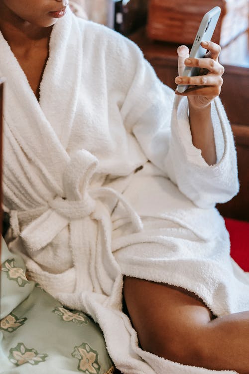 Crop anonymous African American female in bathrobe browsing smartphone while sitting on bed in morning