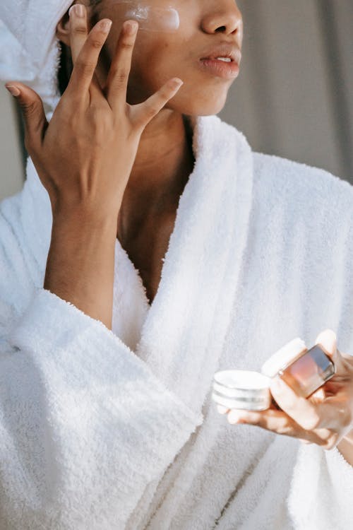 Crop unrecognizable  young female in white bathrobe applying moisturizing cream on face after shower