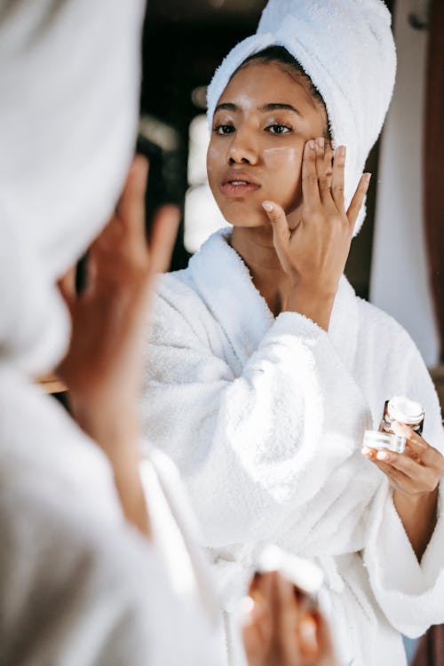 Young female  in white robe and towel on head  applying moisturizing cream on face while standing in bathroom