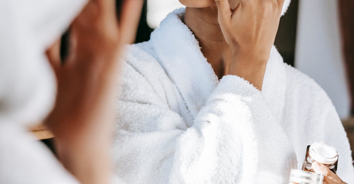 Young black female  in white robe and towel on head  applying moisturizing cream on face while standing in bathroom