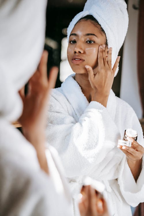 Young black female  in white robe and towel on head  applying moisturizing cream on face while standing in bathroom