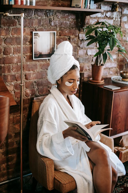 Free Young black female in white bathrobe reading book while sitting on chair  in cozy room Stock Photo