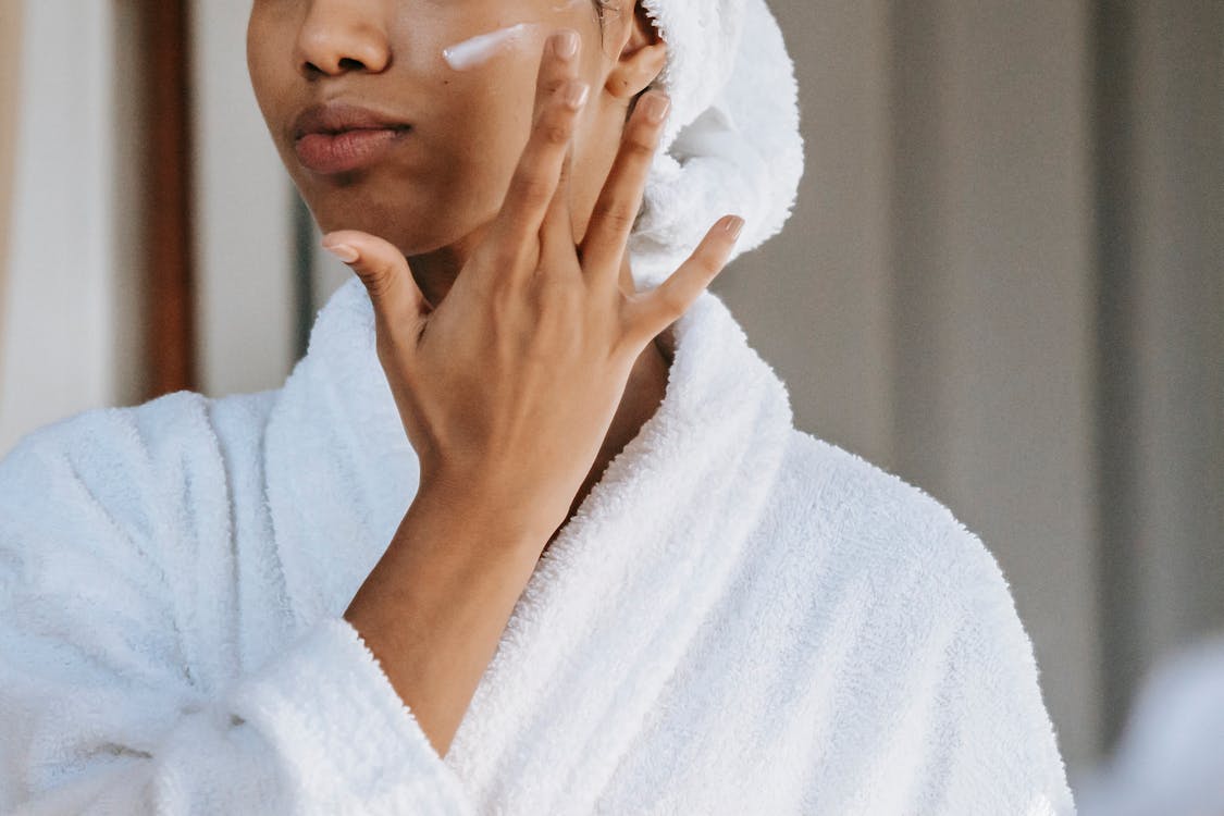 샤워에 서 있는 동안 얼굴에 보습 크림을 바르는 머리에 수건을 얹은 흰색 목욕 가운을 입은 익명의 여성 스톡 사진