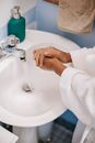 From above of crop anonymous female in white bathrobe washing hands with detergent in sink