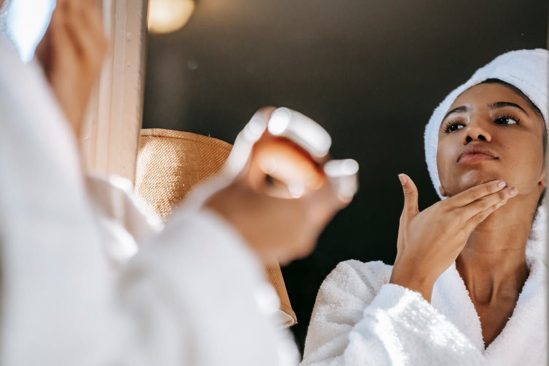 Free African American young female in white bathrobe applying moisturizing cream on face while standing in bathroom Stock Photo