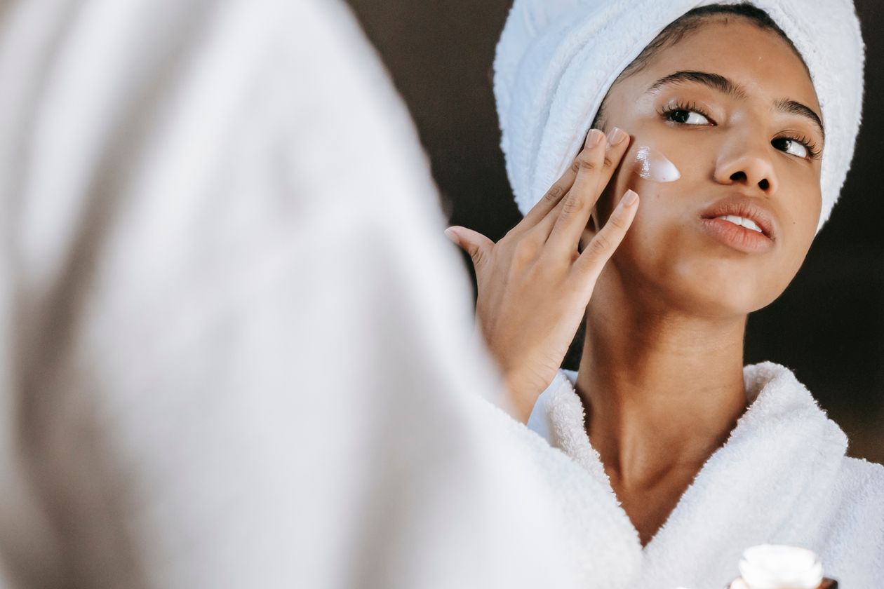 Chica en albornoz aplicándose crema hidratante frente al espejo por la mañana