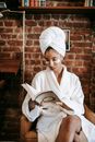 Ethnic female in turban and white robe sitting in armchair and reading book during morning skin care routine
