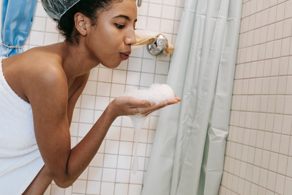 Side view of ethnic female standing in bathroom and blowing foam while washing body during daily hygiene routine