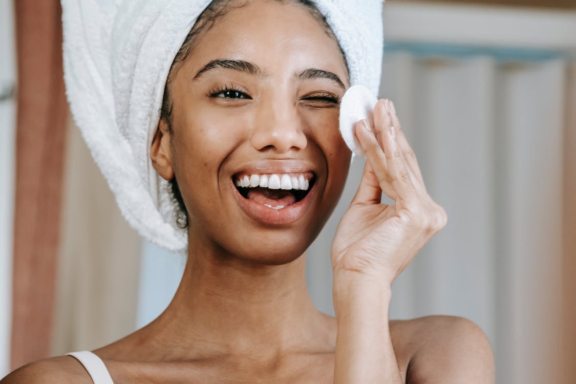 Free Delighted ethnic woman laughing brightly while looking at reflection and cleaning face with cotton pad in morning Stock Photo