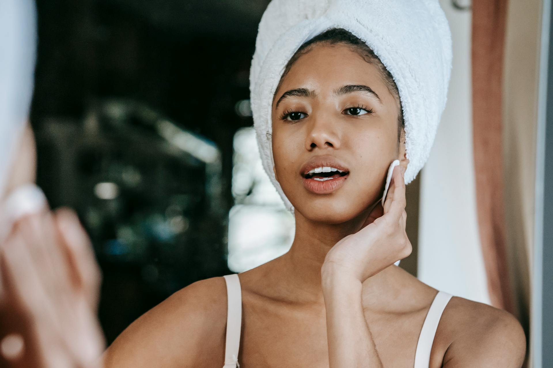 Ethnic female with mouth opened cleaning face with cotton pad while looking at mirror during daily beauty care