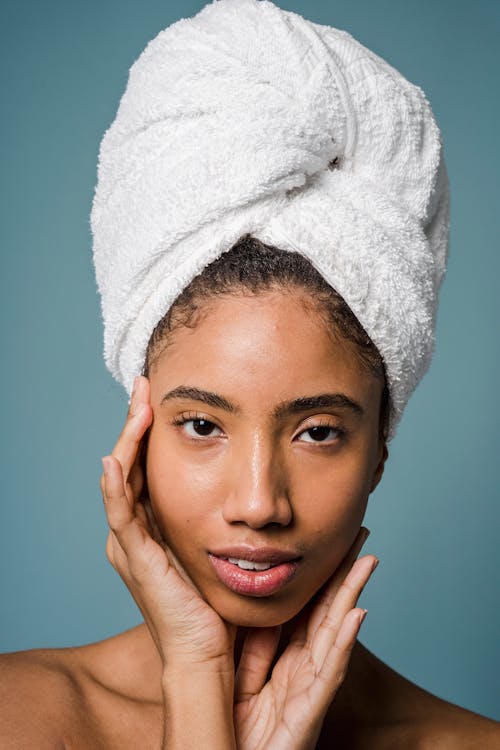 Young African American female with perfect skin and towel on head touching face and looking at camera after shower
