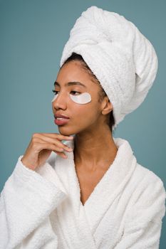 Confident young African American lady with towel on head and eye patches in white bathrobe touching chin and looking away after taking shower