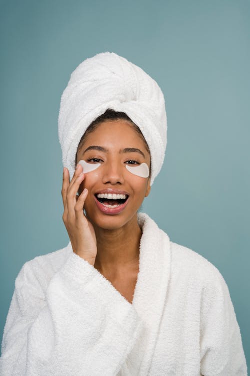 Cheerful young black female in white bathrobe with towel on head and eye patches laughing and looking at camera during skin care routine after shower