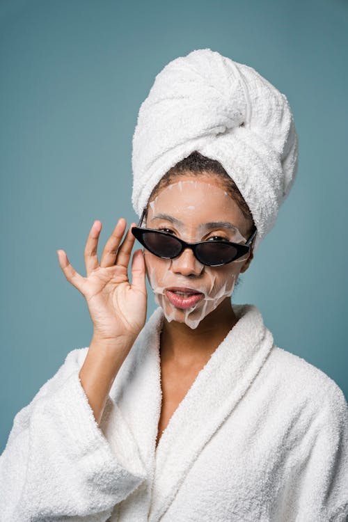 Confident ethnic woman with sheet mask adjusting sunglasses in blue studio