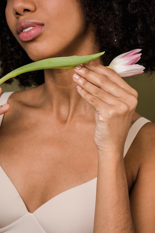 Crop unrecognizable sensual young African American female in stylish lingerie holding delicate tulip flower in hands in studio