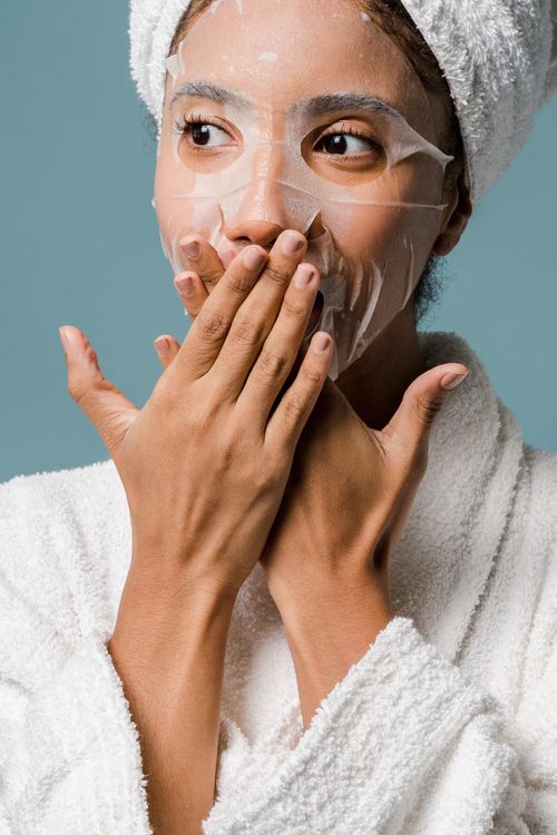 Free Crop surprised young African American female with towel on head and sheet mask in bathrobe covering mouth with hands and looking away during skin care routine against blue background Stock Photo