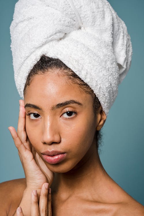 Calm young black lady with perfect skin touching face and looking at camera while standing against blue background with white towel on head after spa procedures