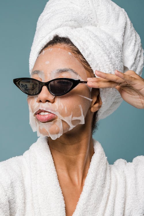 Free Self esteem young African American lady with moisturizing sheet mask and towel on hand wearing trendy sunglasses while standing against blue background in bathrobe after shower Stock Photo