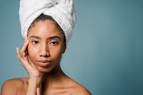 Free Tranquil young black female with pure skin and white towel on head touching face and looking at camera after spa procedures against blue background Stock Photo