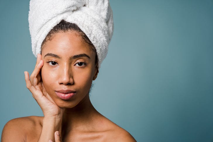 Tranquil young black female with pure skin and white towel on head touching face and looking at camera after spa procedures against blue background