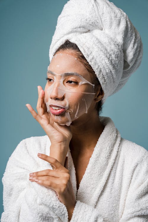 Smiling young ethnic female applying sheet mask on face after shower