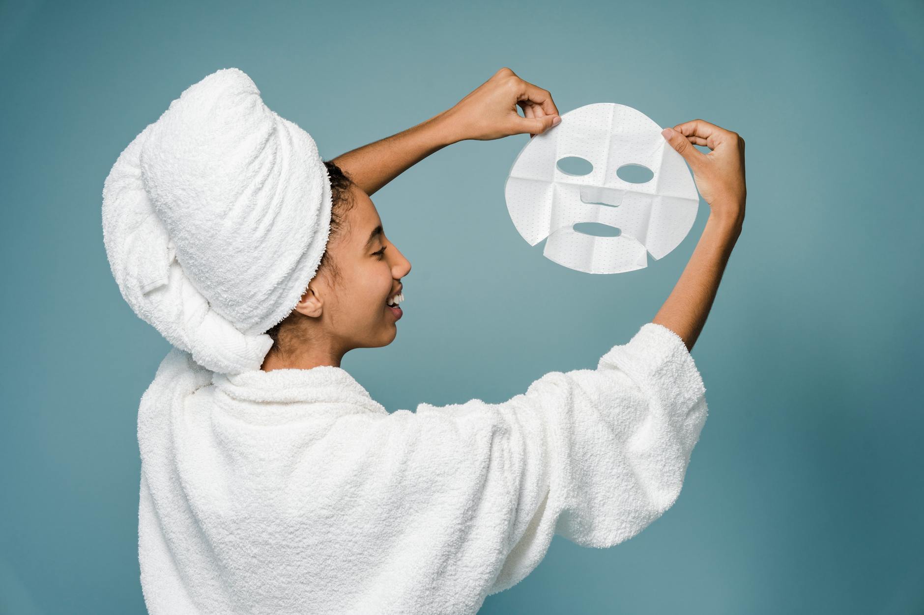 Back view of happy young African American female in bathrobe with towel on head smiling while demonstrating skin care mask against blue background