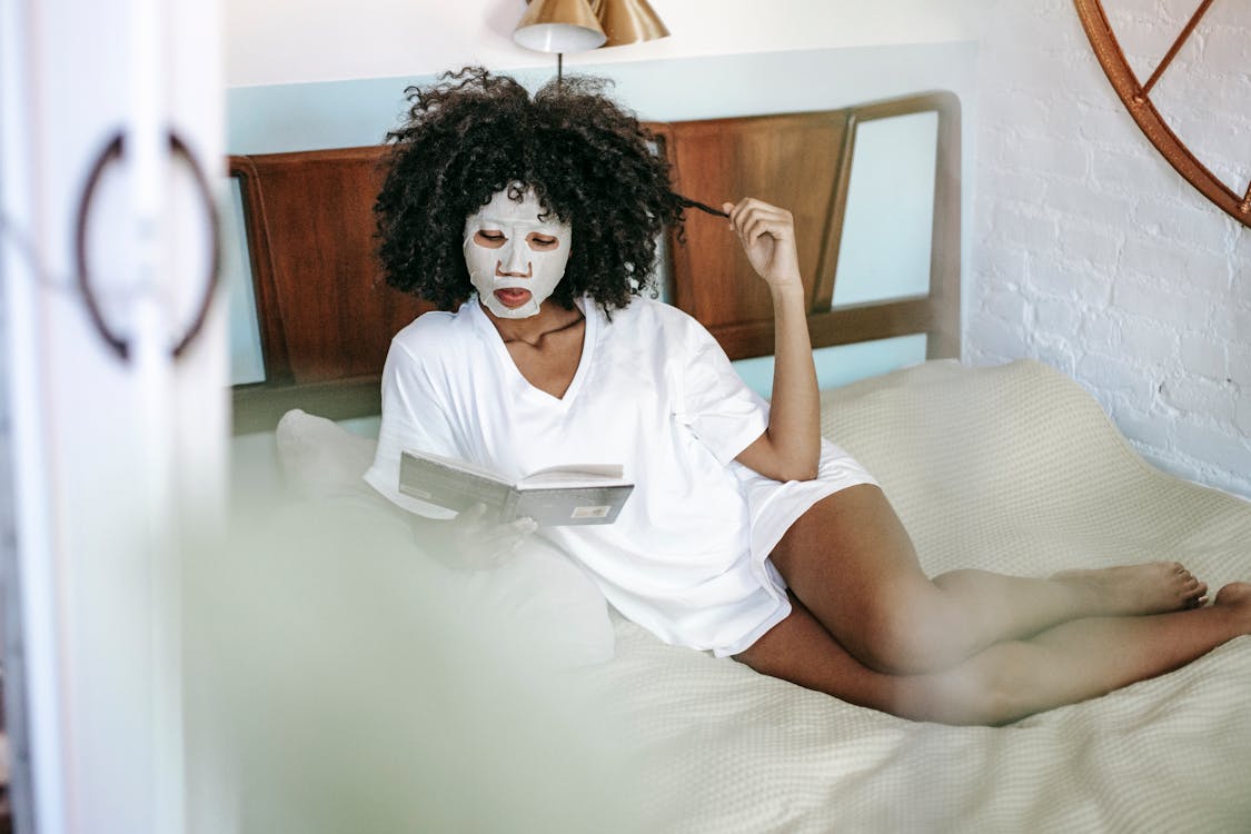 From above of young African American female with sheet mask touching curly hair and reading book while resting on bed at home
