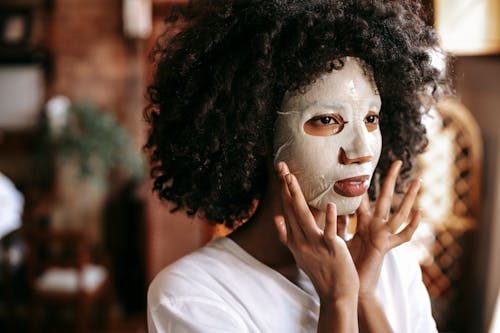 Calm young African American lady with curly hair applying moisturizing sheet mask while relaxing alone at home