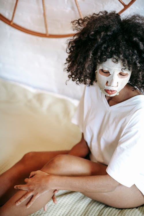 From above of African American woman wearing t shirt sitting on comfortable bed with hydrating sheet mask on face and touching leg