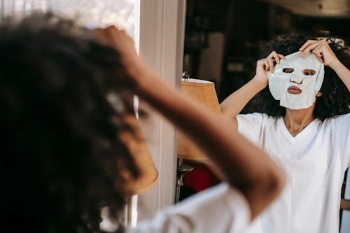Concentrated African American female in domestic cloth putting moisturizing sheet mask while standing near mirror at home in daylight