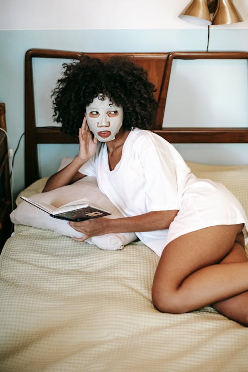 Free High angle of African American woman in white t shirt lying on bed with anti age sheet mask and reading interesting book Stock Photo