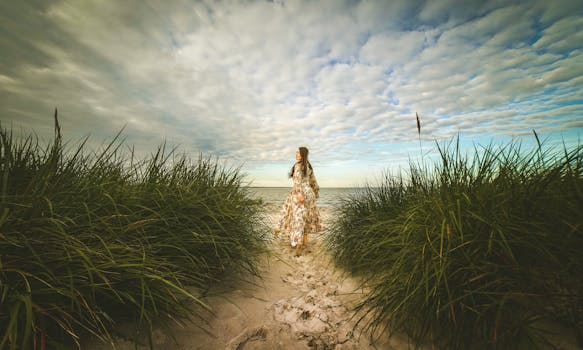 A woman in a floral dress walking on a sandy beach path surrounded by grass under a cloudy sky.