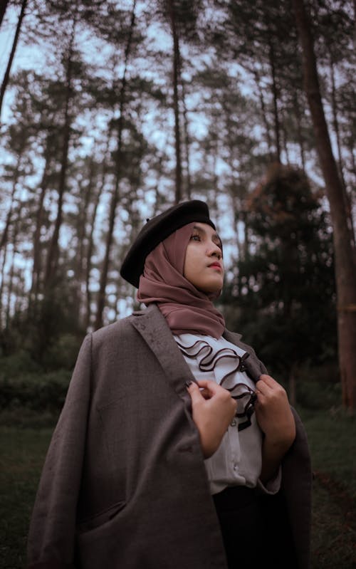 Young Asian female in trendy coat and hat with scarf looking away while standing near trees in woodland