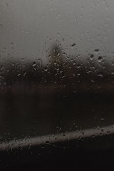 Close-up view of raindrops on a window with a blurred landscape in the background, creating a moody atmosphere.