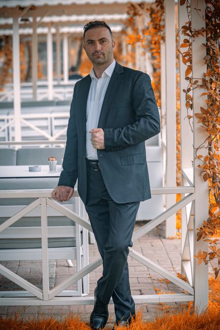 Confident businessman in a suit stands outdoors with autumn foliage.