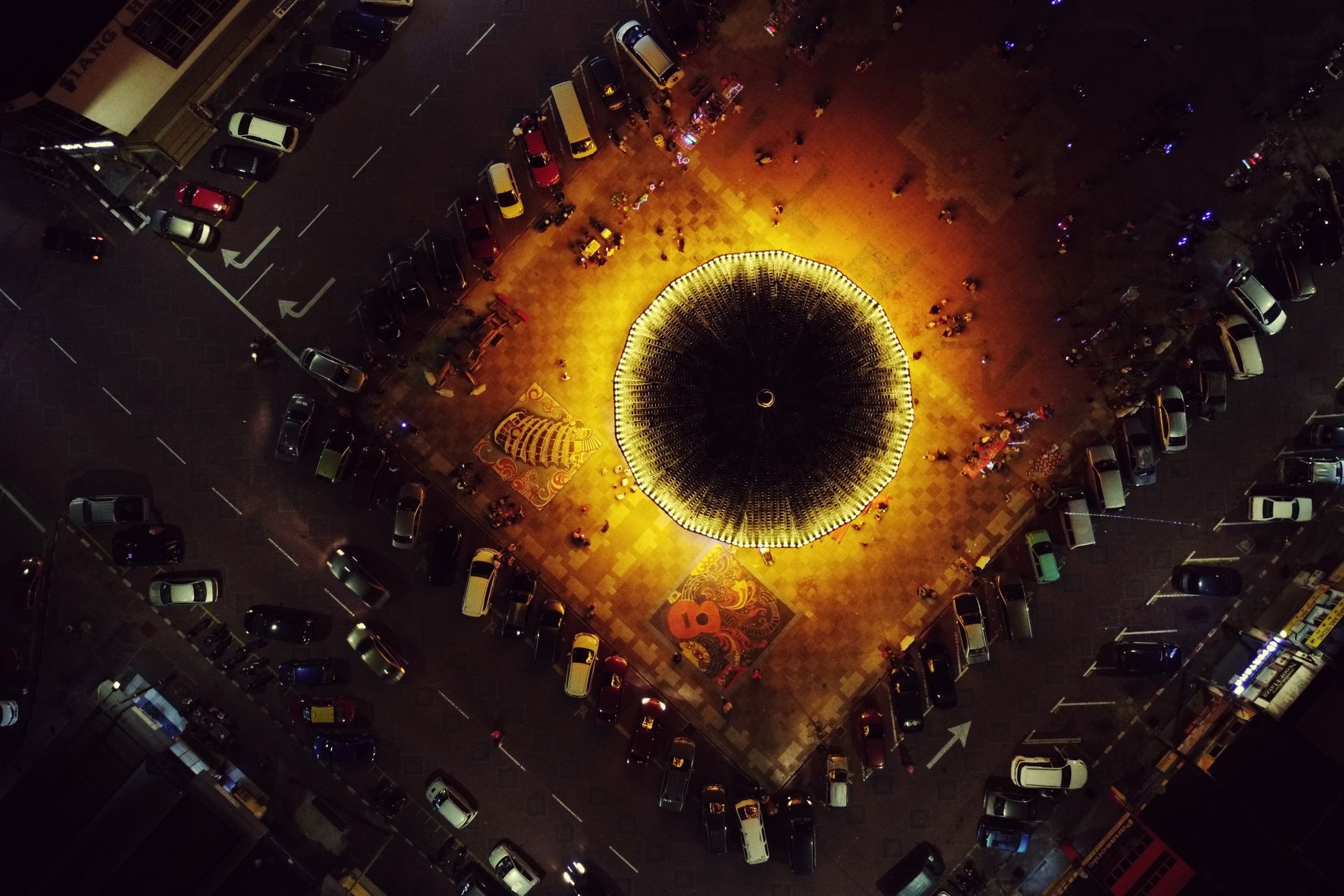 Aerial view of illuminated merry go round on square near road with various parked cars at night