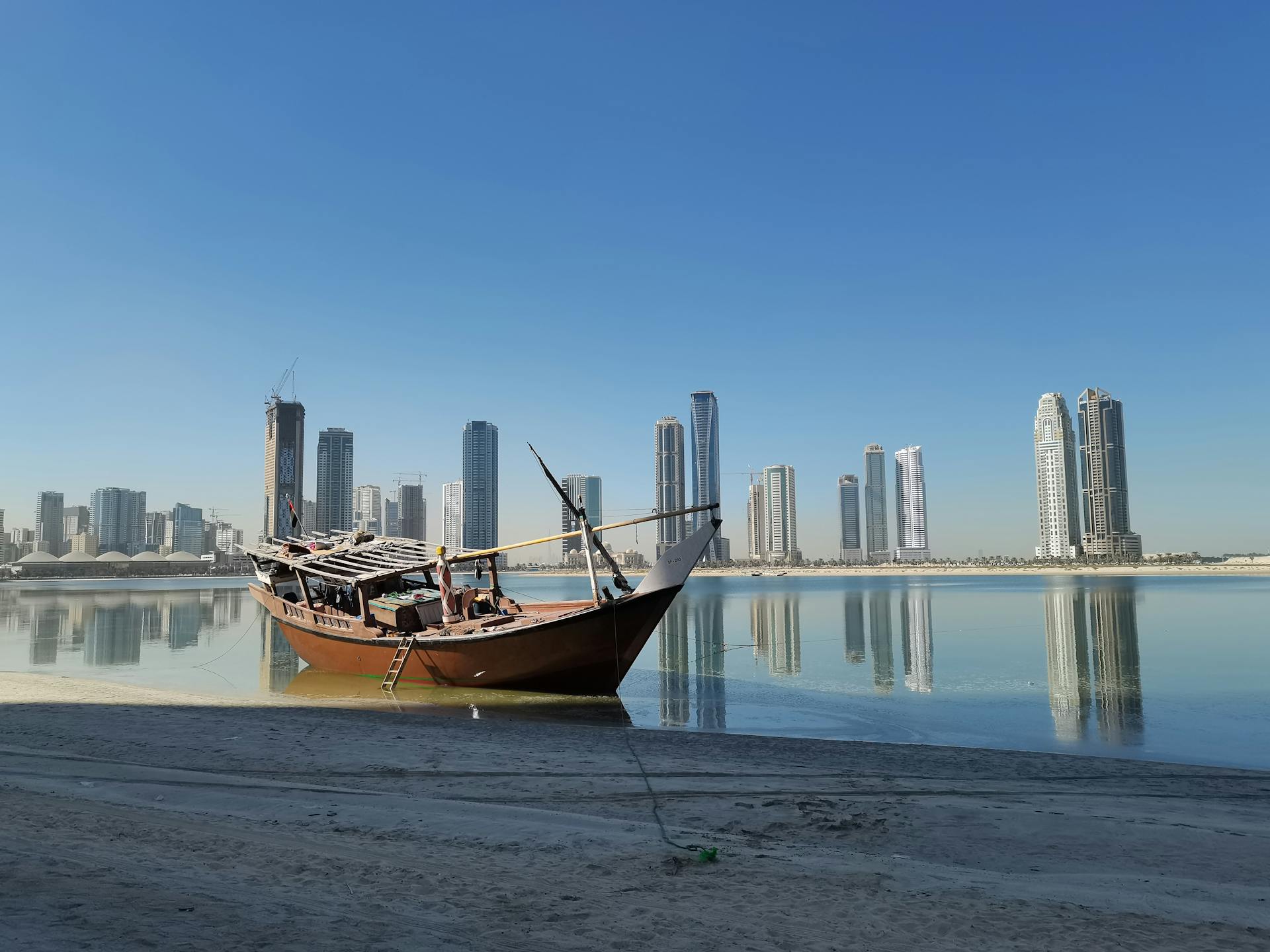 A dhow boat by the Sharjah skyline reflecting in the calm water.
