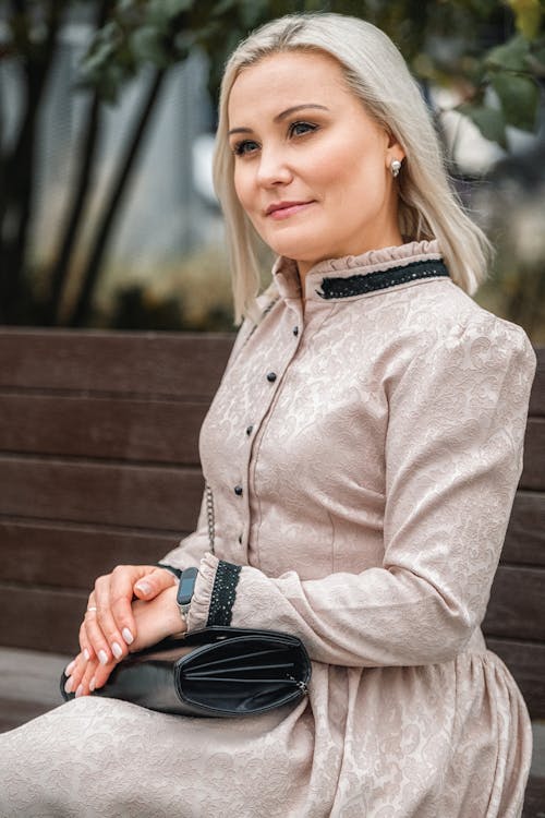 Attractive content female wearing elegant dress sitting on wooden bench in lush city park and looking away in pleasant thoughts