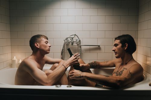 Free A tattooed couple shares an intimate moment, drinking in a warmly lit bathtub. Stock Photo