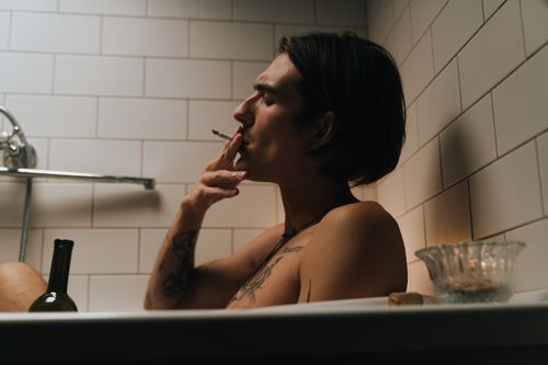 Free A young man relaxes in a white-tiled bathtub, smoking a cigarette with a wine bottle nearby. Stock Photo