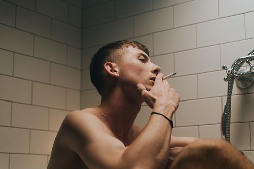 Free Young man in bathroom smoking a cigarette, deep in thought against tiled wall. Stock Photo