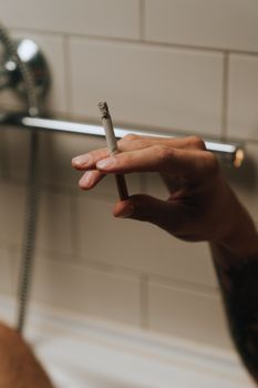 特写镜头：一只手拿着点燃的香烟，靠在浴室瓷砖墙壁上，悬在浴缸上方