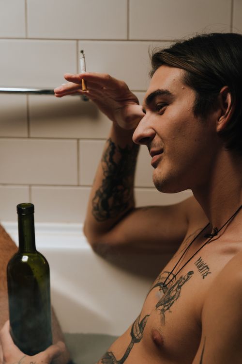 Free A young man with tattoos relaxes in a bathtub, smoking a cigarette, with a wine bottle. Stock Photo